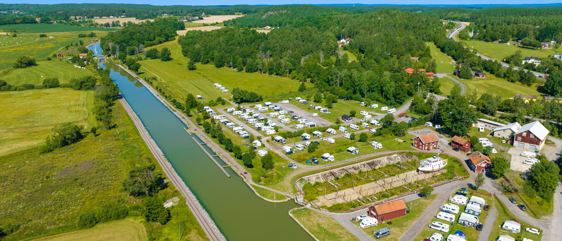 Foto: Flygbild över Göta kanal med Skeppsdockans camping bredvid. Bild visar många husvangnar, husbilar, en torrdocka för båtar samt nröda hus.