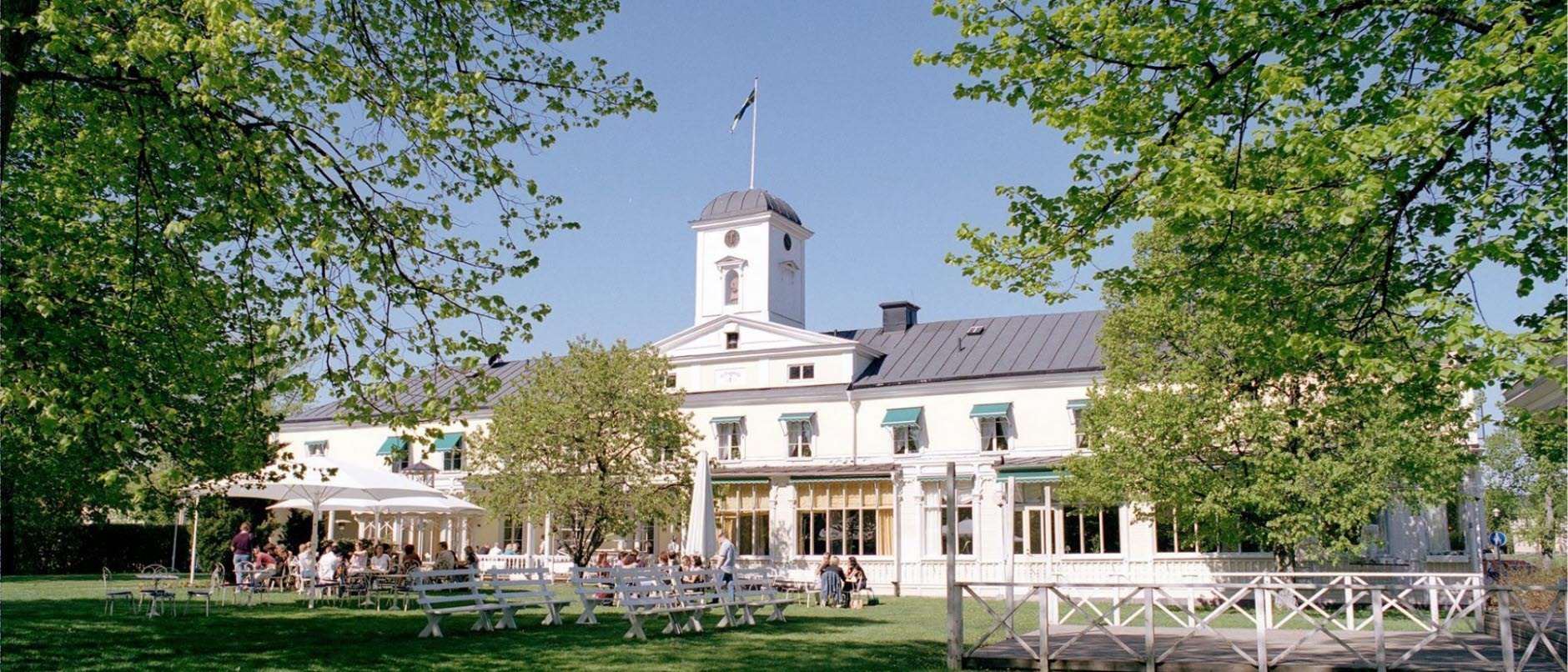 Foto: Bild visar Söderköpings brunns exteriör. En sekelskiftesbyggnad med storvit verande. I förgrunden är det stora lövträd samt en grön gräsmatta och vita cafébord.