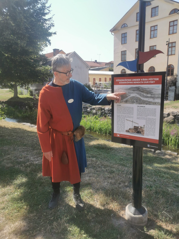 Bild visar en man kl&auml;dd i medeltida kl&auml;der. Han pekar p&aring; en informationstavla