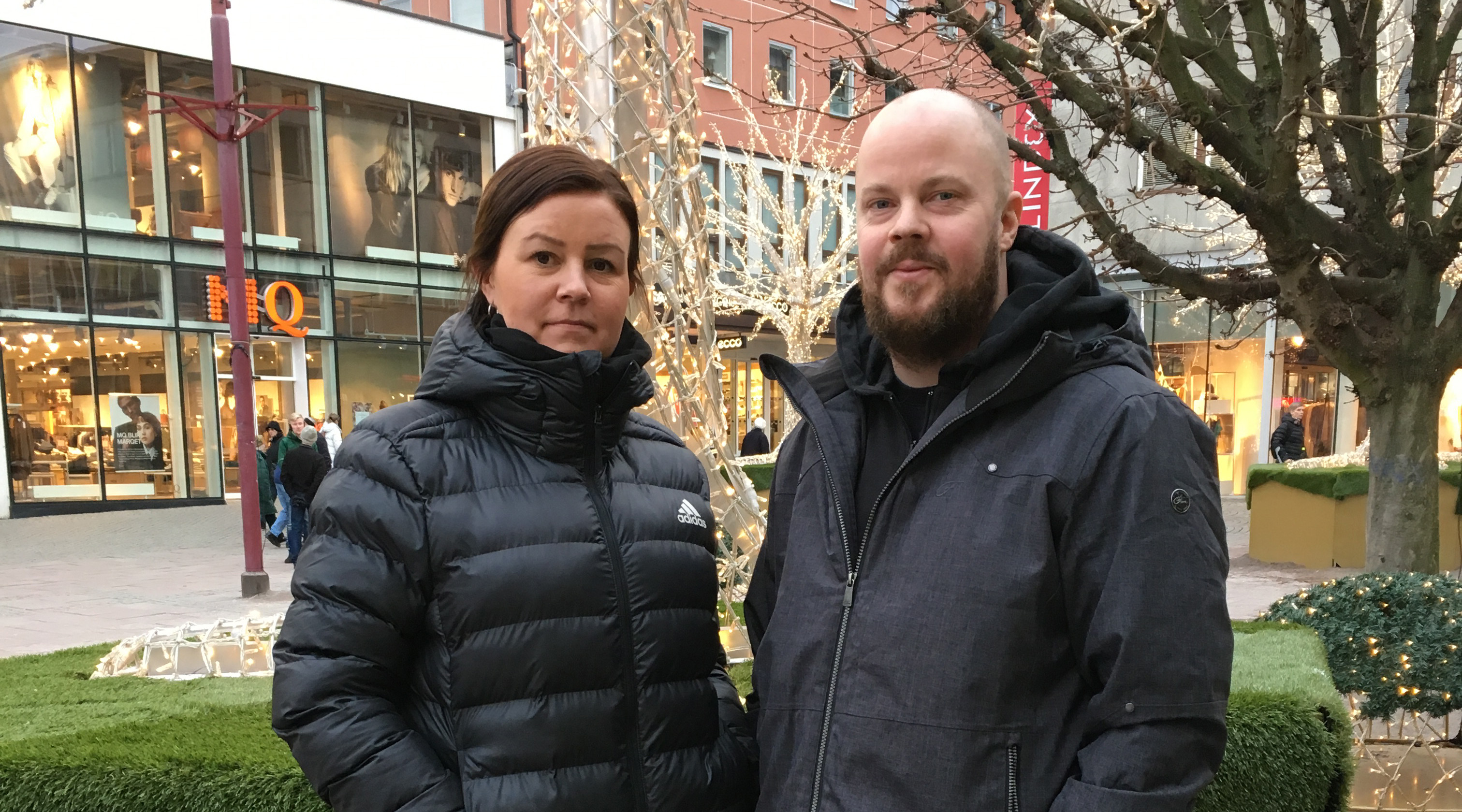 Bilden visar tv&aring; personer som kollar mot kameran. I bakgrunden syns n&aring;gra butiker och tr&auml;d dekorerade med ljusslingor.